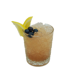 A peach-colored cocktail with crushed ice is served in a patterned short glass and garnished with blueberries and two lemon wedges
