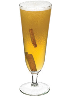 A golden cocktail with a white foamy top is served in a tall stemmed glass with two cinnamon sticks visible inside.