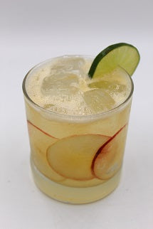 A pale yellow cocktail with ice is served in a short glass with peach slices visible inside and a lime wedge on the rim.