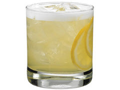 A pale yellow cocktail with ice and a foamy top is served in a short glass with lemon slices visible inside and along the side
