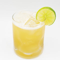 A pale yellow cocktail with ice is served in a short glass and garnished with a lime slice on the rim.