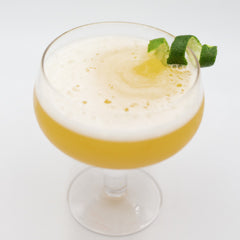 A golden yellow cocktail with a thick white foamy top is served in a coupe glass and garnished with fresh mint leaves and a small yellow citrus garnish.