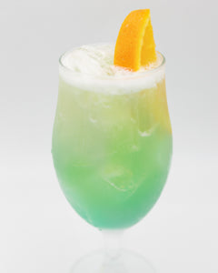 A pale green cocktail with ice is served in a tall stemmed glass with a light foamy top and garnished with an orange peel on the rim.