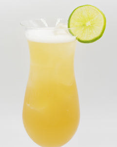 A pale yellow cocktail is served in a tall hurricane glass with a light foamy top and garnished with a lime slice on the rim.