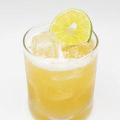 A pale yellow cocktail with ice is served in a short glass and garnished with a lime slice on the rim.