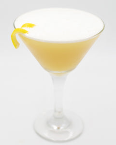 A pale yellow cocktail is served in a coupe glass with a thick white foamy top and garnished with a curled lemon peel on the rim.