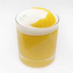 A pale yellow cocktail is served in a short glass with a thick white foamy top and garnished with a curved lemon peel on the surface.