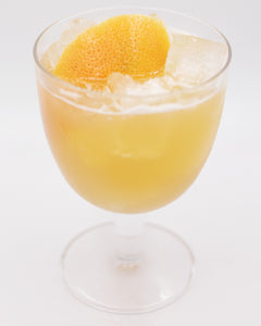 A pale yellow cocktail with ice is served in a rounded stemmed glass and garnished with a large grapefruit peel on top.