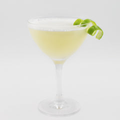 A pale yellow cocktail with a light foamy top is served in a coupe glass and garnished with lime slices and a curled lime peel on the rim.