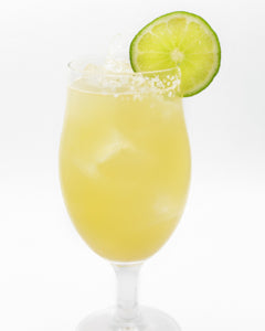 A pale yellow cocktail with ice is served in a tall stemmed glass with a lime slice on the rim and a sugar-coated edge.