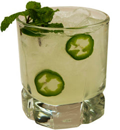 A pale green cocktail with ice is served in a short glass, featuring two jalapeño slices and fresh mint leaves as garnish.