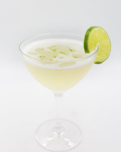 A pale yellow cocktail with a light foamy top is served in a coupe glass, topped with lime zest and garnished with two lime slices on the rim.