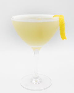 A pale yellow cocktail with a light foamy top is served in a coupe glass and garnished with a long lemon peel on the rim.