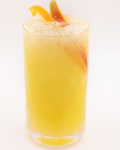 A bright yellow cocktail with ice is served in a tall glass with orange peel pieces visible inside and orange garnishes on top.