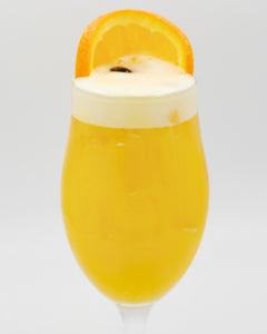 A bright yellow cocktail with a thick white foamy top is served in a tall stemmed glass and garnished with a large orange slice on the rim.