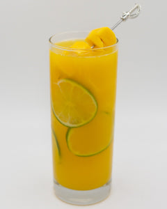 A bright yellow cocktail is served in a tall glass with lime slices visible inside and garnished with mango pieces on a cocktail pick.