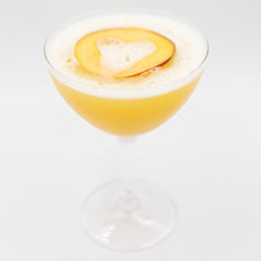 Pale yellow cocktail with foam and peach garnish in a coupe glass