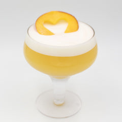 A pale yellow cocktail is served in a coupe glass with a thick white foamy top and garnished with an orange peel arranged on the surface.