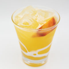 A bright yellow cocktail with ice is served in a short glass with an orange slice visible inside and white decorative swirls around the lower part of the gla