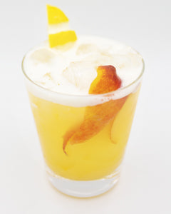 A bright yellow cocktail with ice is served in a short glass with a long orange peel visible inside and small citrus garnishes on top.