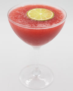 A bright red frozen cocktail is served in a coupe glass with a lime slice floating on the surface.