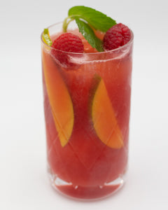 A red cocktail is served in a tall glass with peach slices and raspberries visible inside, garnished with lime slices and fresh mint leaves on top.