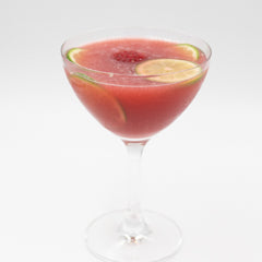 A red cocktail is served in a coupe glass with lime and grapefruit slices floating on the surface.
