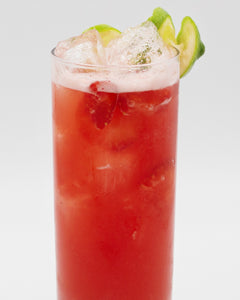 A bright red cocktail with ice is served in a tall glass, topped with a light foamy layer and garnished with lime slices and a lime wedge on the rim.