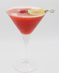 A red cocktail is served in a martini glass with a raspberry and lime slice floating on the surface and a decorative cocktail pick on the rim.