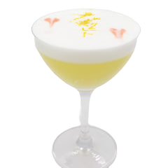 A pale yellow cocktail is served in a coupe glass with a thick white foamy top, decorated with small yellow zest and pink petal-like garnishes.